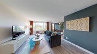 This single-floor residence offers a thoughtfully designed open-concept layout where the living, dining, and kitchen areas flow seamlessly together. Light wood-tone flooring runs throughout, providing a warm, cohesive foundation across all spaces.

The living area is bright and welcoming, anchored by large windows that frame treetop views and flood the room with natural light. Warm rust-colored curtains add a bold contrast to the cool blue accent wall that defines the boundary between the living and dining spaces. A geometric wall panel adds a sculptural touch to the design.

The dining area continues the mid-century modern aesthetic, featuring a teal-painted wall, ceiling fan, and a window that brings in additional natural light. The kitchen is efficiently laid out with grey cabinetry, stone countertops, stainless steel appliances, and a subtle blue tile backsplash.

The bedroom is a calm retreat with its own ceiling fan, a generous window with soft draping curtains, and light-toned walls that create a restful atmosphere. The bathroom features white subway tile wainscoting, a stone-top vanity, and a framed mirror with bright overhead lighting, completing the space with clean, polished finishes.