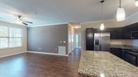 Located at 11211 Westwood Loop in San Antonio, TX, this single-floor unit offers a well-designed open-concept layout that seamlessly connects the living and kitchen areas. The kitchen is a standout feature, showcasing navy cabinetry, a classic white subway tile backsplash with a decorative diamond-pattern wine rack accent, stainless steel appliances including a refrigerator with ice maker, a gas range, and a microwave. A large central island with granite countertops provides additional prep space and casual seating, illuminated by stylish pendant lighting and a track light fixture overhead. Crown molding adds a refined touch throughout the main living areas, which feature rich wood-look flooring. The bedroom areas are carpeted for comfort and include ceiling fans. The primary bedroom connects to a spacious walk-in closet with multi-tier wire shelving and an en-suite bathroom featuring a tub-shower combination. A second full bathroom mirrors similar finishes, including granite countertops and wood-look tile flooring. An additional bedroom benefits from natural light through a window with exterior views. The unit's thoughtful layout and consistent finishes make it a practical and comfortable living space.