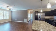 Located at 11211 Westwood Loop in San Antonio, TX, this single-floor unit offers a well-designed open-concept layout that seamlessly connects the living and kitchen areas. The kitchen is a standout feature, showcasing navy cabinetry, a classic white subway tile backsplash with a decorative diamond-pattern wine rack accent, stainless steel appliances including a refrigerator with ice maker, a gas range, and a microwave. A large central island with granite countertops provides additional prep space and casual seating, illuminated by stylish pendant lighting and a track light fixture overhead. Crown molding adds a refined touch throughout the main living areas, which feature rich wood-look flooring. The bedroom areas are carpeted for comfort and include ceiling fans. The primary bedroom connects to a spacious walk-in closet with multi-tier wire shelving and an en-suite bathroom featuring a tub-shower combination. A second full bathroom mirrors similar finishes, including granite countertops and wood-look tile flooring. An additional bedroom benefits from natural light through a window with exterior views. The unit's thoughtful layout and consistent finishes make it a practical and comfortable living space.