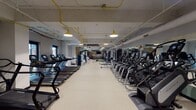 This fitness center is a comprehensively outfitted gym designed to support a wide range of training styles and fitness goals. The space is organized into distinct zones that flow seamlessly from one end to the other, maximizing both functionality and ease of movement.

The cardio zone lines both sides of the main floor with rows of treadmills, elliptical trainers, stair climbers, and stationary bikes — all featuring individual digital displays. The equipment is arranged to allow ample circulation space between rows, contributing to an open and airy feel. Full-length mirrors along one wall visually expand the space and provide form feedback during workouts.

A dedicated strength and functional training area anchors one end of the gym, complete with a turf strip ideal for sled pushes, dumbbell work, and dynamic movements. Adjustable benches, a tiered dumbbell rack, cable machines, and a multi-functional rig with suspension trainers and battle ropes round out this zone. A punching bag and additional functional accessories are also available.

The design aesthetic is modern industrial, with exposed pipework painted in bold yellow running along the ceiling, complemented by matching pendant lighting that casts a warm, focused glow across the floor. A dramatic feature wall displaying the "Hail Hail" text against a nature-inspired mural adds a distinct and motivating character to the space. Light-toned rubberized flooring ties the entire facility together, ensuring durability and comfort underfoot.
