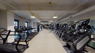 This fitness center is a comprehensively outfitted gym designed to support a wide range of training styles and fitness goals. The space is organized into distinct zones that flow seamlessly from one end to the other, maximizing both functionality and ease of movement.

The cardio zone lines both sides of the main floor with rows of treadmills, elliptical trainers, stair climbers, and stationary bikes — all featuring individual digital displays. The equipment is arranged to allow ample circulation space between rows, contributing to an open and airy feel. Full-length mirrors along one wall visually expand the space and provide form feedback during workouts.

A dedicated strength and functional training area anchors one end of the gym, complete with a turf strip ideal for sled pushes, dumbbell work, and dynamic movements. Adjustable benches, a tiered dumbbell rack, cable machines, and a multi-functional rig with suspension trainers and battle ropes round out this zone. A punching bag and additional functional accessories are also available.

The design aesthetic is modern industrial, with exposed pipework painted in bold yellow running along the ceiling, complemented by matching pendant lighting that casts a warm, focused glow across the floor. A dramatic feature wall displaying the "Hail Hail" text against a nature-inspired mural adds a distinct and motivating character to the space. Light-toned rubberized flooring ties the entire facility together, ensuring durability and comfort underfoot.