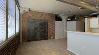 This single-floor space showcases a compelling industrial-loft aesthetic, combining raw architectural elements with practical living or working features. The most striking focal point is a large exposed brick wall, highlighted by a distinctive arched brick detail framing a set of heavy-duty, vintage-style metal vault doors — a nod to the building's historic origins. The brick's warm, textured tones add depth and character to the interior.

Overhead, exposed metal ductwork runs along the ceiling, reinforcing the industrial character of the space, while a pendant light fixture provides a subtle decorative touch. A structural column contributes to the open, loft-like feel of the layout. The flooring is carpeted in a neutral tone, offering a softer contrast to the harder industrial elements throughout.

To one side, a partial wall and countertop — likely a kitchen bar or workspace divider — introduce a functional zone within the open floor plan. White partition walls provide definition without fully enclosing the space, maintaining an open and airy atmosphere. Overall, the space balances historic charm with adaptable, modern utility.