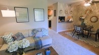 This single-story space features an open-concept layout that seamlessly connects the main living area to the kitchen and dining space. The living room is carpeted and comfortably sized, centered around a plush sofa accented with decorative throw pillows and a cozy blanket. A wooden coffee table anchors the seating area, while framed artwork adorns the neutral-toned walls, adding character to the space. Natural light filters in through a window dressed with vertical blinds, brightening the room with a warm, welcoming ambiance.

A half-wall with a dark countertop pass-through connects the living area to the kitchen, offering both visual openness and functional separation. The kitchen features warm wood cabinetry, a mosaic tile backsplash, and dark appliances, providing a practical yet stylish cooking space. Decorative wall art is displayed above the pass-through counter, adding a personalized touch.

The dining area, positioned adjacent to the living room, accommodates casual seating and features tile flooring, creating a natural transition between the carpeted lounge space and the kitchen. Overall, the space offers a cohesive, comfortable layout with warm tones throughout.