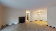 Located at 7433 Old Sauk Rd in Madison, Wisconsin, this single-floor residence offers a thoughtfully laid-out living space with a clean, neutral aesthetic throughout. The open living area serves as the heart of the home, anchored by a classic wood-mantled fireplace with a glass-front surround and complemented by plush carpet underfoot. A pendant light fixture illuminates the adjacent dining area, which transitions seamlessly into the kitchen via hardwood-style flooring. The galley-style kitchen is well-appointed with rich, dark wood cabinetry, granite countertops, a deep basin sink, and stainless steel appliances including a range, microwave, and refrigerator — all set against a bright, efficiently organized layout. Bedrooms are carpeted and generously sized, with one featuring a large sliding-glass closet door and a window overlooking mature trees, bringing in pleasant natural light and greenery. The bathroom includes a full-size soaking tub and shower combination with clean, white surroundings. Throughout the space, baseboard heating, dark wood trim, and neutral white walls create a cohesive and functional interior suited for comfortable everyday living.