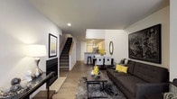 This three-floor space masterfully combines contemporary residential design with industrial architectural elements. The main living level features an open-concept layout where the living, dining, and kitchen areas flow seamlessly into one another. The living room centers around a sleek fireplace and is styled with modern furnishings, hardwood-style flooring, and crisp white walls accented with bold décor. A staircase with natural wood detailing connects the floors, while recessed lighting keeps the space bright and inviting. The kitchen and dining area are visible in the background, offering a functional and well-appointed layout with bar seating and a defined dining space.

An entry-level area showcases dramatic floor-to-ceiling windows framed by exposed brick columns and timber beam accents, flooding the space with natural light and providing views of a landscaped courtyard with bicycle storage — a nod to the building's industrial heritage.

An upper-level common or workspace area further highlights the industrial aesthetic, with soaring ceilings, exposed ductwork, and aged brick walls. A communal high-top table with bar stools sits along a mezzanine-style railing, creating an open, collaborative atmosphere enhanced by pendant lighting and abundant vertical space.