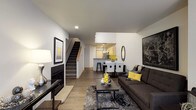 This three-floor space masterfully combines contemporary residential design with industrial architectural elements. The main living level features an open-concept layout where the living, dining, and kitchen areas flow seamlessly into one another. The living room centers around a sleek fireplace and is styled with modern furnishings, hardwood-style flooring, and crisp white walls accented with bold décor. A staircase with natural wood detailing connects the floors, while recessed lighting keeps the space bright and inviting. The kitchen and dining area are visible in the background, offering a functional and well-appointed layout with bar seating and a defined dining space.

An entry-level area showcases dramatic floor-to-ceiling windows framed by exposed brick columns and timber beam accents, flooding the space with natural light and providing views of a landscaped courtyard with bicycle storage — a nod to the building's industrial heritage.

An upper-level common or workspace area further highlights the industrial aesthetic, with soaring ceilings, exposed ductwork, and aged brick walls. A communal high-top table with bar stools sits along a mezzanine-style railing, creating an open, collaborative atmosphere enhanced by pendant lighting and abundant vertical space.