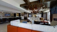 This single-floor space showcases a thoughtfully designed interior that blends industrial and contemporary aesthetics. The most striking architectural feature is the exposed dark wood beam ceiling installation, from which clusters of bare Edison-style bulbs hang on vivid red cords, creating a dramatic and artistic lighting focal point that anchors the entire space.

The lounge area is generously proportioned, featuring multiple distinct seating arrangements. Large, low-profile sofas in neutral gray tones are paired with bold orange leather accent seating, creating a dynamic contrast that energizes the space. Coffee tables and side tables provide functional surfaces throughout the conversational groupings, while a blue ottoman adds a playful pop of color.

Dark wood paneling lines portions of the walls, complementing the ceiling beams and adding warmth to the space. A large world map graphic adorns one wall, contributing to a cosmopolitan atmosphere. In the background, a bar or counter area with high stools is visible, suggesting a social beverage station.

Large mirrors and glass elements enhance the sense of openness, while the light-toned flooring provides a clean contrast to the darker wood accents. The overall ambiance is modern, inviting, and well-curated.