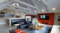 This single-floor space makes a strong first impression with its dramatic vaulted ceiling, defined by crisp white-painted exposed beams and trusses that lend an airy, open quality to the interior. Skylights and track lighting are integrated throughout the ceiling structure, ensuring the space is well-illuminated at all times of day. The main lounge area is anchored by a sleek, tufted black leather sofa and matching armchairs arranged around a rustic wooden coffee table with hairpin legs, creating an inviting seating arrangement ideal for socializing or relaxing. A bold burnt-orange accent wall serves as a striking focal point, housing a large wall-mounted flat-screen television and built-in cabinetry below. Complementing this are coordinating red-orange accent furnishings that reinforce the vibrant color palette throughout. Tall decorative grasses in a centerpiece vase add a natural, organic touch to the contemporary setting. In the background, a reception or service counter is visible, suggesting a shared amenity or community-oriented function for the space. The flooring transitions between warm hardwood and patterned carpet, further delineating functional zones within the open layout. Overall, the space balances modern design with a welcoming, energetic atmosphere.