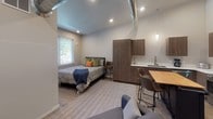 Located at 821 E Mullan Ave in Coeur d'Alene, Idaho, this modern single-floor studio offers a thoughtfully designed open-plan layout that seamlessly integrates living, sleeping, dining, and cooking areas into one cohesive space. Soaring ceilings with exposed industrial-style ductwork lend a loft-like character, while light wood flooring and a neutral color palette create a clean, contemporary aesthetic throughout.

The kitchen is well-appointed with rich wood cabinetry, white quartz countertops, stainless steel appliances including a full-size refrigerator, built-in microwave, and range. A central island with a butcher-block top and seating for two provides an ideal dining and prep area, anchored by a sleek pendant light above.

The sleeping area is positioned to take advantage of natural light from multiple windows fitted with blinds, offering views of the surrounding greenery. A tall wood wardrobe cabinet provides ample storage, while a built-in nook with a bench and sliding barn-style mirror door near the living area adds rustic warmth and additional organization.

The living area features a comfortable sectional sofa and connects fluidly to the rest of the space. A full bathroom with modern fixtures completes the unit. A frosted-glass entry door allows light while maintaining privacy, contributing to the bright and welcoming ambiance of this well-designed studio.
