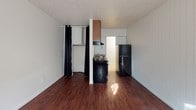 Located at 18 N Main St in Brockport, NY, this single-floor unit offers a straightforward and functional layout designed for efficient everyday living. The main living area is finished with rich, dark hardwood floors that extend throughout the space, complemented by white walls and a textured ceiling that adds subtle character. A front entry door features a decorative arched transom window, allowing natural light to filter in alongside a side window dressed with curtain panels. A wall-mounted air conditioning unit provides climate control directly in the living area.

The kitchen is positioned toward the rear of the unit and is outfitted with warm wood cabinetry, dark laminate countertops, a stainless steel sink, a black electric coil-burner range with an overhead ventilation hood, and a black refrigerator. A curtained closet area adjacent to the kitchen provides additional storage space.

The bathroom is clean and efficiently arranged, featuring light tile flooring, a single vanity with a mirror, and a full tub-and-shower combination with white tile surround. A grab bar and towel rod are also present, adding practical touches to the space.

Overall, this unit presents a no-frills, move-in-ready layout in a convenient Brockport location, ideal for those seeking a compact and functional living space.
