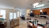 This single-floor residence offers an open-concept design that integrates the living, dining, and kitchen areas into one cohesive and functional space. The living room is bright and inviting, featuring large windows with blinds that allow generous natural light to fill the room, complemented by a ceiling fan overhead and light-toned tile flooring that extends throughout the common areas. Crown molding adds a refined architectural detail along the ceiling, enhancing the overall sense of polish and quality.

The dining area is situated adjacent to the kitchen, anchored by a classic round wooden dining table and chairs, and illuminated by an elegant pendant light fixture. The kitchen features warm honey-toned cabinetry, dark granite-style countertops, and a breakfast bar that serves as a natural transition between the cooking and dining spaces. Additional pendant lighting above the kitchen area adds both function and style. A full-size refrigerator and ample upper cabinet storage round out the kitchen's practical offerings.

Throughout the space, the neutral color palette of soft beiges and warm whites creates a calm, welcoming ambiance. The open layout and thoughtful placement of lighting fixtures contribute to a comfortable, well-lit environment that feels both spacious and easy to navigate.