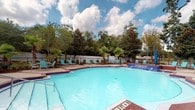 Located at 2220 Gloria Cir in Pensacola, Florida, this single-level outdoor space centers around a generously sized swimming pool that serves as its primary feature. The pool's clear, light-blue water is complemented by blue-tiled accents along the pool's edge, giving it a clean, resort-style aesthetic. The pool area accommodates a range of depths, making it suitable for various users.

Surrounding the pool is a well-maintained deck furnished with turquoise lounge chairs and small accent tables, providing ample space for relaxation and sunbathing. The deck surface features a warm, earth-toned finish that pairs naturally with the outdoor setting.

The perimeter of the space is enclosed by a black iron fence, ensuring both security and an open, airy feel. Beyond the fencing, a lush backdrop of mature trees — including tall magnolias and tropical palm trees — frames the area and provides natural shade and privacy, creating a serene, garden-like atmosphere.

The overall ambiance of the space is bright and inviting, enhanced by the natural Florida landscape and the open sky above. Whether used for leisure or recreation, this pool area offers a comfortable and visually appealing outdoor retreat in a well-established Pensacola neighborhood.