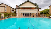 This outdoor amenity space centers around a generously sized rectangular swimming pool with clearly marked lane dividers, offering ample room for both lap swimming and leisure. The pool's vivid blue water is complemented by a wide concrete sundeck that surrounds it on all sides, furnished with numerous lounge chairs and umbrella-shaded seating areas — ideal for relaxing outdoors.

The two-story clubhouse building anchors one end of the pool area and serves as the architectural focal point of the space. The ground level features large glass doors that open directly to the pool deck, providing easy indoor-outdoor access. Ceiling fans are installed on both levels of the covered porch, ensuring comfort during warm weather. The upper level offers an open-air covered veranda with additional ceiling fans and views overlooking the pool area.

The pool deck is enclosed by a black iron fence, providing both security and a clear boundary. Mature trees are visible beyond the fence line, offering a natural backdrop and partial shade. The surrounding multi-story residential buildings frame the space, creating an intimate community atmosphere. Safety equipment, including a life ring, is visibly mounted near the pool entrance, reflecting attention to safety standards throughout the space.