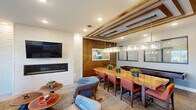 Located at 2511 Watermark Ter in Elgin, IL, this thoughtfully designed community lounge space combines a living room and dining area into a single, cohesive floor plan. The interior showcases a contemporary aesthetic, blending warm wood accents with bold, vibrant design elements throughout.

The living area is anchored by a teal velvet loveseat accented with decorative throw pillows, positioned on a neutral area rug over light hardwood flooring. A wall-mounted flat-screen TV sits above a sleek linear gas fireplace framed by white shiplap, creating a striking focal point. An adjacent wet bar features textured mosaic tile from floor to ceiling, under-counter beverage refrigeration, and pendant lighting in turquoise, adding a lively pop of color. Large sliding glass doors flood the space with natural light and provide direct access to an outdoor patio area.

The dining section is defined by a stunning live-edge wood communal table paired with orange upholstered chairs, illuminated by a substantial glass-and-metal rectangular pendant light fixture. Decorative wood beams accent the ceiling above, adding architectural warmth and dimension. A shuffleboard table along the wall offers a recreational amenity, while interior windows connect the space visually to adjoining areas. A vibrant teal wood-panel accent wall further enlivens the dining zone, making this lounge an inviting and multifunctional gathering space.