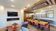 Located at 2511 Watermark Ter in Elgin, IL, this thoughtfully designed community lounge space combines a living room and dining area into a single, cohesive floor plan. The interior showcases a contemporary aesthetic, blending warm wood accents with bold, vibrant design elements throughout.

The living area is anchored by a teal velvet loveseat accented with decorative throw pillows, positioned on a neutral area rug over light hardwood flooring. A wall-mounted flat-screen TV sits above a sleek linear gas fireplace framed by white shiplap, creating a striking focal point. An adjacent wet bar features textured mosaic tile from floor to ceiling, under-counter beverage refrigeration, and pendant lighting in turquoise, adding a lively pop of color. Large sliding glass doors flood the space with natural light and provide direct access to an outdoor patio area.

The dining section is defined by a stunning live-edge wood communal table paired with orange upholstered chairs, illuminated by a substantial glass-and-metal rectangular pendant light fixture. Decorative wood beams accent the ceiling above, adding architectural warmth and dimension. A shuffleboard table along the wall offers a recreational amenity, while interior windows connect the space visually to adjoining areas. A vibrant teal wood-panel accent wall further enlivens the dining zone, making this lounge an inviting and multifunctional gathering space.