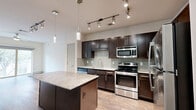 This thoughtfully designed one-bedroom, one-bathroom unit offers a functional and modern layout across a single floor. The open-concept kitchen and living area serves as the heart of the unit, showcasing rich dark espresso cabinetry, a sage green subway tile backsplash, and a full suite of stainless steel appliances including a refrigerator, range, microwave, and dishwasher. A granite-topped center island provides additional prep space and casual seating, while stylish pendant lights and track lighting illuminate the space. Durable wood-look flooring runs throughout the kitchen and living areas, complementing the warm, contemporary aesthetic. The living room is bright and airy, featuring a ceiling fan and a sliding glass door that opens to a private balcony with tree-lined views, allowing abundant natural light to fill the room. The bedroom is carpeted for comfort, with a large window offering additional natural light and pleasant outdoor views. Two closets provide generous storage, including a walk-in closet with built-in shelving. The bathroom features clean white tile flooring, a soaking tub with shower, and direct access to the walk-in closet. Additional conveniences include a dedicated laundry area, pantry, and utility room, making this a complete and practical living space.