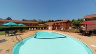 This outdoor pool area serves as a central recreational amenity, showcasing a generously sized freeform swimming pool with clear, blue-green water. The pool is framed by a wide paved deck that provides ample space for relaxation, furnished with multiple lounge chairs and an umbrella for shade. Safety features, including a life ring station, are conveniently positioned poolside. The surrounding landscape includes mature trees that provide a natural backdrop, enhancing the open-air atmosphere under clear skies.

Adjacent to the pool area, the property features single-story brick buildings with a classic, well-maintained exterior. Interior spaces include carpeted rooms finished with neutral-toned walls and clean white trim and paneled doors. The rooms are equipped with wall-mounted air conditioning units and windows fitted with blinds that allow natural light to filter in while offering privacy. Select rooms include walk-in closets, adding practical storage. A carpeted hallway connects the rooms, maintaining a cohesive and comfortable interior aesthetic throughout. Together, the outdoor pool and interior spaces offer a well-rounded living environment that balances leisure and function.