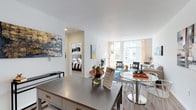 This thoughtfully designed single-floor residence offers a seamless open-concept layout that connects the kitchen, dining, and living areas into one cohesive, light-filled space. The kitchen is outfitted with stainless steel appliances — including a refrigerator, range, microwave, and dishwasher — complemented by white cabinetry, a subway tile backsplash, light countertops, track lighting, and durable hardwood-style flooring. A mobile stainless steel island adds flexible prep and storage space. The open dining area flows naturally into the living room, which benefits from floor-to-ceiling windows that flood the space with natural light and provide views of the surrounding greenery. The living room features clean white walls, hardwood-style flooring, and a modern aesthetic. Two bedrooms are carpeted for comfort; the primary bedroom includes a walk-in closet, while the second bedroom features a window with outdoor views. The bathroom offers a clean, minimalist design with a white vanity, undermount sink, modern fixtures, and a large mirror. A dedicated laundry area houses a stacked washer and dryer unit alongside wire shelving, adding everyday convenience to this well-appointed residence.