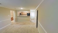 This single-floor space offers an open and functional layout that seamlessly connects the living area to the kitchen. The living space is finished with neutral beige carpet flooring and warm-toned walls, creating a calm and welcoming ambiance throughout. Crown molding and clean white baseboards add a touch of classic detail to the interior.

The kitchen features light oak cabinetry with ample storage and is accessible via a pass-through counter that opens directly to the living area, promoting an open feel between spaces. Black appliances, including what appears to be a microwave and refrigerator, are integrated into the cabinetry. Natural light filters in through a window near what appears to be an entryway door at the rear of the kitchen area, brightening the space.

A doorway visible on the right side of the living area leads to additional rooms, suggesting a practical flow throughout the floor plan. The consistent use of neutral colors across walls, flooring, and trim creates a cohesive, understated aesthetic that is both versatile and easy to personalize.