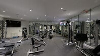 This fitness center is a fully outfitted exercise facility designed to support a wide range of workout routines. The space is laid out across a single floor and is divided into distinct functional zones, each dedicated to a specific type of training. The main workout area features floor-to-ceiling mirrors lining the walls, visually expanding the room and allowing users to monitor their form. Recessed lighting throughout the ceiling provides consistent, bright illumination across the entire space. The flooring is a durable rubber gym surface, offering both traction and impact absorption. The strength training zone is well-stocked with a variety of cable machines, weight machines, and multi-functional cable stations, alongside a full rack of dumbbells ranging across multiple weight increments and kettlebells. A dedicated cardio area houses upright and recumbent stationary bikes as well as treadmills. Mounted flat-screen televisions are positioned throughout for entertainment during workouts. A storage area with built-in cubbies provides space for personal belongings, while a small refrigeration unit is available for beverages. The overall aesthetic is clean and contemporary, with neutral grey and white tones creating a focused and professional atmosphere.