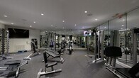 This fitness center is a fully outfitted exercise facility designed to support a wide range of workout routines. The space is laid out across a single floor and is divided into distinct functional zones, each dedicated to a specific type of training. The main workout area features floor-to-ceiling mirrors lining the walls, visually expanding the room and allowing users to monitor their form. Recessed lighting throughout the ceiling provides consistent, bright illumination across the entire space. The flooring is a durable rubber gym surface, offering both traction and impact absorption. The strength training zone is well-stocked with a variety of cable machines, weight machines, and multi-functional cable stations, alongside a full rack of dumbbells ranging across multiple weight increments and kettlebells. A dedicated cardio area houses upright and recumbent stationary bikes as well as treadmills. Mounted flat-screen televisions are positioned throughout for entertainment during workouts. A storage area with built-in cubbies provides space for personal belongings, while a small refrigeration unit is available for beverages. The overall aesthetic is clean and contemporary, with neutral grey and white tones creating a focused and professional atmosphere.