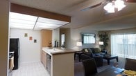 This single-story space is designed with an open-concept floor plan that seamlessly connects the kitchen and living area, creating a cohesive and functional environment. The kitchen features light wood cabinetry, a built-in dishwasher, and a peninsula with an integrated sink that doubles as a breakfast bar, providing both workspace and a natural dividing point between the two areas. Overhead, a recessed drop-panel light fixture brightens the kitchen, while the textured ceiling adds a classic architectural detail throughout. The living area is carpeted and comfortably furnished with dark leather seating, a coffee table, and floor lamps that contribute to a warm, inviting ambiance. A large framed mirror on the wall enhances the sense of depth, while vertical blinds on the window allow for adjustable natural light. A sizable indoor plant adds a touch of greenery to the living space. The neutral color palette of warm beige and tan tones is consistent throughout, contributing to a unified and understated aesthetic. The layout promotes easy flow between functional zones, making the space well-suited for both everyday living and casual entertaining.