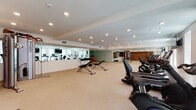 Located at 22520 Lyons Ave in Santa Clarita, CA, this single-level fitness center is thoughtfully designed to support a wide range of exercise disciplines. The space is organized into distinct functional zones, creating an efficient and professional gym environment.

The main workout area features a generous selection of cardio equipment, including multiple treadmills and stationary bikes, each equipped with individual display screens for interactive workouts. Wall-mounted flat-screen televisions are positioned throughout for added convenience. A dedicated free weights section includes an adjustable weight bench and a dumbbell rack, suitable for strength training routines.

Floor-to-ceiling mirrors line the walls throughout the facility, visually expanding the space and allowing users to monitor their form during exercise. A separate barre studio area features hardwood-style flooring and wall-mounted barre rails alongside its own mirrored wall, making it ideal for dance fitness, yoga, or stretching sessions.

The facility is finished with neutral-toned carpeting in the main workout zone and is illuminated by recessed ceiling lighting, creating a clean, bright, and functional atmosphere. Accent brick wall elements add a subtle design contrast to the otherwise crisp white interior. Natural light filters in through multiple windows, further enhancing the open and inviting environment.