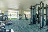 Located at Bixby Village in Long Beach, California, this single-story fitness center offers a bright, modern environment designed to support a comprehensive workout experience. The interior is finished with distinctive teal-toned carpet flooring that adds a vibrant, contemporary feel to the space. Large windows along multiple walls flood the room with natural light and provide views of the surrounding landscaping and community buildings. A full wall of mirrors enhances the sense of openness throughout the workout area. The fitness center is thoroughly equipped with a multi-station cable and strength training machine, a free weight rack with a full range of dumbbells, an adjustable weight bench, stationary cycling bikes with individual display screens, elliptical cross-trainers, and treadmills. Wide glass doors open directly onto a covered outdoor transition area with a slatted wood pergola and synthetic turf, seamlessly connecting the fitness center to the resort-style outdoor pool deck. The pool area features ample lounge seating, market umbrellas, and a clean concrete surround enclosed by a white fence. The overall ambiance of the fitness center is clean, airy, and inviting, making it a well-rounded amenity space suited for a variety of fitness routines.
