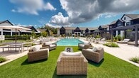 Located at 20 Graf Dairy Drive in Mobile, Alabama, this thoughtfully designed residential community showcases craftsman-style architecture with dark board-and-batten siding, gabled rooflines, and large windows that lend the buildings a refined, residential character. The centerpiece of the outdoor amenity space is a generously sized resort-style swimming pool surrounded by travertine pool decking and an abundance of lounge chairs, offering ample space for relaxation under the Alabama sun. Adjacent to the pool, lush green lawn areas are furnished with wicker seating arrangements and a fire pit, creating an inviting social atmosphere. A striking open-air pavilion with exposed timber framing anchors one end of the courtyard, providing shaded outdoor dining and lounge space complete with wood-topped tables and comfortable seating. Herringbone brick pathways wind through the property, connecting various outdoor areas and adding a classic, warm aesthetic to the grounds. Meticulously maintained landscaping, including ornamental shrubs, native plantings, and manicured garden beds, frames the entire outdoor space. The community's gated entrance is framed by white brick columns and black iron fencing, reinforcing a sense of security and curb appeal throughout the property.