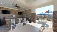 This outdoor lounge and clubhouse facility offers a well-designed social environment that seamlessly blends indoor comfort with outdoor living. The covered patio area is anchored by a full brick accent wall fitted with a wall-mounted flat-screen TV and a built-in bar counter lined with white high-back bar stools, ideal for casual gatherings. A ceiling fan overhead provides airflow, making the shaded space comfortable year-round. Open-air dining tables and chairs extend the seating area, while views of the surrounding pool deck — complete with lounge chairs and large market umbrellas — create a resort-like atmosphere.

The interior clubhouse portion features soaring vaulted ceilings with recessed lighting and a statement ceiling fan. Polished concrete floors run throughout, reflecting natural light that floods in through expansive floor-to-ceiling windows overlooking the pool. A stone-surround fireplace with a mounted TV serves as the focal point of the main lounge area, flanked by comfortable seating and tasteful artwork. A sleek bar counter with dark granite and warm wood cabinetry, illuminated by pendant lights, provides additional gathering space. The open layout, contemporary furnishings, and abundant natural light combine to create an inviting and functional community space.