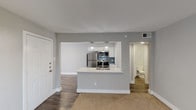 This single-floor residence is designed with a clean, modern aesthetic highlighted by neutral gray walls, white trim, and a consistent mix of wood-style plank flooring and plush carpet throughout. The open-concept layout connects the living area directly to the kitchen via a peninsula counter with a white surface and integrated sink, creating a functional and social hub at the heart of the unit. The kitchen is well-equipped with stainless steel appliances—including a refrigerator, electric range with microwave above, and dishwasher—complemented by white shaker-style cabinetry, a blue tile backsplash, and pendant lighting above the peninsula. A separate entry door provides direct access to the main living area, which features a vertical blind-covered sliding glass door that invites natural light. The bedrooms are carpeted and include ceiling fans with light fixtures, windows dressed with horizontal blinds, and ample wall space. A hallway connects the private areas of the unit, with a full bathroom featuring a vanity, toilet, and white finishes. An additional closet is accessible from the hallway, adding practical storage. Overall, the space presents a tidy, updated interior suited for comfortable everyday living.