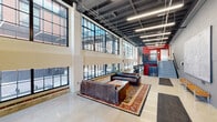 Elton Park is a striking development located in Detroit's historic Corktown neighborhood, blending industrial heritage with modern design. The lobby serves as the centerpiece of the building, welcoming visitors through a set of tall glass-and-steel entry doors beneath a soaring double-height ceiling. A bold branded entry mat announces the space's identity immediately upon arrival.

The main lobby area is defined by its dramatic two-story steel-framed warehouse windows, which flood the interior with abundant natural light and offer views of the surrounding urban streetscape. The exposed black structural ceiling grid reinforces the building's industrial character, while polished concrete floors provide a clean, contemporary contrast. A seating area anchored by a leather sofa arrangement on a richly patterned area rug creates a comfortable, inviting gathering space within the expansive interior.

A secondary corridor area features a large architectural map of Detroit mounted prominently on a grey concrete block wall, accompanied by wooden benches — a nod to the building's deep civic and neighborhood roots. Industrial-style linear lighting runs throughout, ensuring the space is well-illuminated at all times. A steel staircase with mesh railings, accented with bold red architectural elements, connects to adjacent levels and adds visual dynamism. The overall ambiance is one of purposeful, urban sophistication rooted in Detroit's industrial legacy.