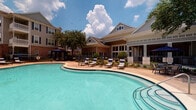 Located at 2402 N Ben Wilson St in Victoria, Texas, this outdoor amenity space centers around a generously sized, freeform swimming pool with striking turquoise water. The pool area is designed for comfort and leisure, offering an expansive concrete sundeck lined with cushioned lounge chairs and striped accent pillows, ideal for relaxing under the Texas sun. A dedicated spa is positioned adjacent to the main pool, providing a more intimate retreat. Shaded cabanas with navy blue draped curtains offer a private lounging experience, each furnished with reclining chairs and overlooking the surrounding landscaped grounds. The meticulously maintained landscaping features ornamental shrubs, native plants, rock-bed gardens, and mature trees, all enclosed within decorative wrought-iron fencing that defines the pool courtyard. The space is framed by multi-story brick and siding buildings featuring balconies, dormer windows, and classic architectural detailing. A clubhouse-style structure with large windows anchors one side of the courtyard, enhancing the community feel of the space. The overall ambiance is bright, open, and resort-inspired, making it a well-appointed outdoor destination suited for both relaxation and social gathering.