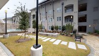 This single-story shared laundry facility is part of a larger multi-story residential complex distinguished by its stucco exterior, arched architectural detailing, and private balconies. The surrounding grounds feature a well-maintained courtyard with a manicured lawn, ornamental trees, low shrubs, and a series of evenly spaced concrete stepping stones that guide residents along defined pathways to the facility entrance.

Inside, the laundry room is defined by its industrial-influenced design, showcasing exposed steel roof beams, visible ductwork, and soaring ceilings that contribute a sense of openness. The polished concrete floors are durable and easy to maintain, reinforcing the utilitarian character of the space. Natural light filters in through windows near the entrance door, supplemented by overhead fluorescent lighting that ensures consistent illumination throughout.

A long row of white commercial-grade washing machines lines one wall, with a corresponding row of dryers mounted above or positioned adjacent. A dark countertop surface runs along one side, providing a practical folding and sorting area. An adjacent waiting area includes seating along the walls, offering residents a comfortable place to wait. The layout is straightforward and functional, prioritizing ease of use and efficient circulation throughout the space.