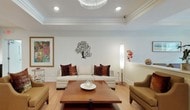 This single-floor space is thoughtfully designed to offer both comfort and functionality across several distinct areas. The lounge areas are finished with warm, neutral tones and light hardwood flooring throughout, creating a cohesive and inviting atmosphere. Tray ceilings with crown molding add architectural depth, while recessed lighting and a statement crystal chandelier enhance the sense of refinement. Seating areas are furnished with tan upholstered armchairs and sofas accented by rich, jewel-toned throw pillows. Decorative touches—including framed artwork, a tall potted plant, and curated accessories—contribute to a polished, welcoming environment.

The kitchenette is outfitted with rich espresso cabinetry, white quartz countertops, stainless steel appliances including a large French door refrigerator and built-in microwave, and glass-front upper cabinets. Under-cabinet lighting highlights the clean lines of the workspace, which also features a stainless steel sink. A corridor from the kitchenette leads directly into a dedicated fitness room, visible through a glass-paneled door and equipped with exercise balls and open floor space. The overall design balances professionalism with warmth.