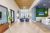 Located at 12175 Philips Hwy in Jacksonville, FL, this thoughtfully designed single-floor clubhouse offers a generous and versatile community space. The grand entrance immediately impresses with soaring ceilings adorned by exposed wood beam detailing and a striking sputnik chandelier, all set above herringbone-patterned light wood flooring that flows throughout the main area.

The open lounge features multiple seating arrangements with plush navy blue accent chairs and a living area anchored by a sleek linear fireplace set against a floor-to-ceiling white marble surround, complemented by large windows that flood the space with natural light and outdoor views of palm trees. A bold navy feature wall with geometric paneling frames a large flat-screen TV and shuffleboard table in the adjacent game room area.

A built-in kitchenette with an island, bar seating, open shelving, and modern appliances provides a functional social hub. A living moss accent wall and curated artwork add organic texture throughout. Three private offices, accessed through a corridor lined with black-framed glass partitions, offer quiet workspaces — each furnished with a dedicated desk or meeting table, artistic lighting, and patterned carpet tiles — ideal for focused work or small meetings.