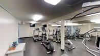 This fitness center is designed to support a full cardio-focused workout routine in a clean, functional setting. The space is outfitted with multiple treadmills, elliptical trainers, and stationary bikes, providing a well-rounded selection of cardiovascular equipment for users of varying fitness levels. A stability ball and additional accessories complement the machine lineup. The flooring features durable, rubber-style material suited for high-traffic athletic use, while the neutral-toned walls and structural columns contribute to the room's straightforward, utilitarian aesthetic. Full-length mirrors line one wall, enhancing the sense of openness and allowing users to monitor their form during workouts. Overhead flush-mount lighting panels illuminate the entire space evenly, ensuring visibility throughout. Wall-mounted flat-screen televisions are positioned for easy viewing from the cardio equipment, offering entertainment during workouts. The single-floor layout keeps everything accessible and organized within one open room, making navigation simple and efficient. Overall, this fitness center provides a practical and well-maintained environment equipped with the essentials for an effective exercise routine.
