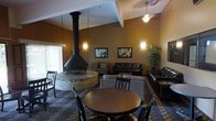 This single-floor space is a well-appointed lounge or common area designed for comfort and social gathering. The room's most striking architectural feature is a dramatic freestanding conical fireplace set atop a circular brick hearth at the center of the space, serving as a natural focal point and gathering spot. Soaring vaulted ceilings with exposed beam accents add a sense of openness and volume, while pendant and wall-mounted lighting cast a warm, ambient glow throughout. The space is furnished with a mix of leather sofas and upholstered seating arranged thoughtfully to encourage conversation, complemented by a round wooden dining table and chairs for more structured gatherings. A patterned blue area rug anchors the seating arrangements and adds a layer of visual interest to the flooring. Warm earth-toned walls adorned with framed artwork and a lush potted plant contribute to the welcoming atmosphere. Large windows along one side allow natural light to filter in, brightening the interior. The overall ambiance is cozy yet spacious, blending rustic elements like the brick fireplace with contemporary furnishings to create an inviting, functional communal environment.