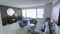 This single-floor space presents a thoughtfully arranged living area defined by its clean, contemporary design. Large windows spanning much of the exterior wall flood the room with natural light, complemented by horizontal blinds that allow for adjustable privacy and light control. The neutral wall tones — a soft grey accent wall contrasting with lighter surrounding surfaces — lend the space a modern, understated aesthetic.

The living area is furnished with a pair of plush, blue-grey barrel chairs accented with decorative pillows, arranged around a sleek glass-top coffee table. A coordinating sofa anchors one side of the seating arrangement, reinforcing the cohesive colour scheme. Soft, light-toned carpeting runs throughout, contributing to the warm and comfortable ambiance.

A compact dining area is positioned near the windows, featuring a round bistro-style table with slim metal chairs, making efficient use of the natural light available. Along one wall, a dark console with a round mirror above it adds a stylish, functional element to the space. A potted plant near the window introduces a natural, organic touch to the overall interior.