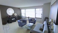 This single-floor space presents a thoughtfully arranged living area defined by its clean, contemporary design. Large windows spanning much of the exterior wall flood the room with natural light, complemented by horizontal blinds that allow for adjustable privacy and light control. The neutral wall tones — a soft grey accent wall contrasting with lighter surrounding surfaces — lend the space a modern, understated aesthetic.

The living area is furnished with a pair of plush, blue-grey barrel chairs accented with decorative pillows, arranged around a sleek glass-top coffee table. A coordinating sofa anchors one side of the seating arrangement, reinforcing the cohesive colour scheme. Soft, light-toned carpeting runs throughout, contributing to the warm and comfortable ambiance.

A compact dining area is positioned near the windows, featuring a round bistro-style table with slim metal chairs, making efficient use of the natural light available. Along one wall, a dark console with a round mirror above it adds a stylish, functional element to the space. A potted plant near the window introduces a natural, organic touch to the overall interior.