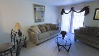 This single-floor residential space offers a warm and inviting atmosphere anchored by a thoughtfully arranged living area. The living room serves as the heart of the space, featuring soft, plush wall-to-wall carpeting that adds a sense of comfort underfoot. A large sliding glass door or wide window with decorative valance treatments allows an abundance of natural light to fill the room, creating a bright and airy ambiance throughout the day. The living area is centered around a plush sofa accented with patterned throw pillows, complemented by a sleek, round glass-topped coffee table with a dark metal frame that adds a contemporary touch. An ornate, decorative mirror with a mosaic-style frame serves as a standout wall accent, adding visual depth and character to the neutral-toned walls. The overall color palette is soft and neutral, with warm beige and gray tones creating a cohesive and calming environment. The layout is practical and well-proportioned, offering an efficient and functional flow. The space is well-suited for comfortable everyday living, combining classic furnishing choices with subtle decorative elements that give the interior a polished, move-in-ready feel.