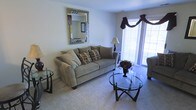 This single-floor residential space offers a warm and inviting atmosphere anchored by a thoughtfully arranged living area. The living room serves as the heart of the space, featuring soft, plush wall-to-wall carpeting that adds a sense of comfort underfoot. A large sliding glass door or wide window with decorative valance treatments allows an abundance of natural light to fill the room, creating a bright and airy ambiance throughout the day. The living area is centered around a plush sofa accented with patterned throw pillows, complemented by a sleek, round glass-topped coffee table with a dark metal frame that adds a contemporary touch. An ornate, decorative mirror with a mosaic-style frame serves as a standout wall accent, adding visual depth and character to the neutral-toned walls. The overall color palette is soft and neutral, with warm beige and gray tones creating a cohesive and calming environment. The layout is practical and well-proportioned, offering an efficient and functional flow. The space is well-suited for comfortable everyday living, combining classic furnishing choices with subtle decorative elements that give the interior a polished, move-in-ready feel.