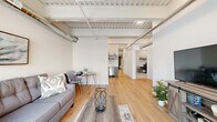 Located at 101 North Queen Street in the heart of downtown Lancaster, PA, this modern urban loft blends industrial character with contemporary finishes throughout its thoughtfully designed single-floor layout. The open-concept kitchen and living area serves as the centerpiece of the space, showcasing light hardwood flooring, soaring ceilings with exposed steel beams, corrugated metal deck, and visible ductwork that give the unit a distinctive loft aesthetic. Large windows in the living room flood the space with natural light and offer views of the surrounding cityscape. The kitchen features sleek gray cabinetry, stainless steel appliances including a refrigerator and dishwasher, white countertops, and open shelving. A dedicated office provides a private, carpeted workspace beneath the dramatic exposed ceiling. The bedroom is a quiet, carpeted retreat with a large window overlooking the urban streetscape. The full bathroom is finished with rich dark wood cabinetry, a white countertop vanity, and neutral tile flooring. A utility room with an in-unit washer adds everyday convenience. Two hallways connect the spaces efficiently, and a closet provides dedicated storage. The overall ambiance is modern and industrial yet warm and livable.