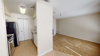 This single-floor residence offers a practical and comfortable layout designed for everyday living. The kitchen is well-appointed with white cabinetry, light granite-style countertops, and stainless steel appliances including an electric range, over-the-range microwave, and a black refrigerator. Light wood-look flooring runs through the kitchen and dining areas, providing a clean, easy-to-maintain surface. A partial wall divides the kitchen from the adjoining spaces, creating defined zones while maintaining an open feel. The living area is generously sized, featuring warm neutral carpet, built-in wall shelving, and a fireplace, making it a versatile and inviting space. A ceiling fan with a light fixture adds comfort and airflow to the dining area, which connects seamlessly to the kitchen. The bedroom is a quiet, carpeted retreat with a window fitted with blinds that allows natural light to filter in. A closet with white bifold doors offers convenient storage. The bathroom is clean and functional, complete with wood-look flooring, a vanity with granite-style countertop, a full tub and shower combination with a curved curtain rod, and crisp white tile surround. Neutral beige tones and textured ceilings are consistent throughout, giving the space a cohesive, understated aesthetic.