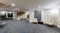 Located at 31 W Patrick St in historic Frederick, Maryland, this single-floor commercial space offers a generously sized and thoughtfully appointed layout. The centerpiece of the space is a grand reception and lounge area distinguished by an ornate coffered ceiling with detailed crown molding, recessed lighting, and decorative wall reliefs. Patterned blue-toned carpeting flows throughout, complementing the neutral color palette of the walls. Comfortable seating arrangements with upholstered sofas and lounge chairs create a welcoming atmosphere, while tall windows allow natural light to filter in. A classic wooden staircase with a polished banister serves as an architectural focal point, anchored by a white column base that adds to the refined, traditional character of the space. Corridors are accented with slate-blue wall panels and framed artwork, lending a professional yet sophisticated ambiance. A dedicated private office features built-in wraparound desk surfaces, a drop ceiling with fluorescent lighting, and the same coordinating blue-accented lower wall detailing found throughout the space. This cohesive design makes the space well-suited for professional or client-facing business use in the heart of downtown Frederick.