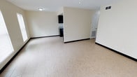 This single-floor space presents an open and functional layout designed for comfortable everyday living. The main living area is expansive and airy, finished with large light-toned ceramic floor tiles that run throughout and contribute to a bright, clean aesthetic. Dark baseboards provide a subtle contrast against the neutral cream-colored walls, adding a touch of definition to the space.

A ceiling fan with an integrated light fixture is centrally positioned in the living area, ensuring efficient air circulation and supplemental lighting. Recessed vents are visible along the walls and ceiling, indicating a built-in HVAC system for year-round climate control.

A partial dividing wall separates the living area from the kitchen zone, which features dark cabinetry and a countertop with a sink, visible from the main space. This semi-open configuration allows for connectivity between the two areas while maintaining a degree of separation. A white interior door leads to an additional room or utility area, rounding out the accessible portions of the floor plan.

Overall, the space offers a straightforward, well-maintained interior that is move-in ready, with consistent finishes and a neutral palette that provides a versatile foundation for a variety of uses and furnishing styles.
