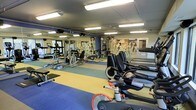 This fitness center is designed to accommodate a full spectrum of workout needs within a single, efficiently organized floor. The space is divided into distinct functional zones, identifiable by its multi-colored rubber flooring — featuring blue, yellow, and gray sections — which helps delineate areas for different types of exercise and provides both cushioning and traction underfoot.

The cardio zone is well-stocked with stationary bikes, including a recumbent cycle, alongside treadmills and elliptical machines, offering members a variety of low-impact and high-intensity options. The strength training area features a selection of cable machines, weight benches, and multi-function resistance equipment, catering to both targeted muscle training and full-body workouts.

Full-length mirrors line the walls, enhancing the sense of space while allowing users to monitor their form during exercise. Overhead fluorescent lighting ensures the entire area is brightly and evenly lit. Decorative frosted glass panels add a refined visual element, softening the utilitarian feel of the space.

The layout is thoughtfully arranged to allow ample circulation between equipment, promoting a safe and comfortable training environment. A separate room visible through a glass door suggests additional dedicated space, potentially for stretching or group fitness activities.