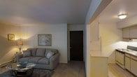 This single-floor residential unit offers a practical and well-organized layout designed for comfortable everyday living. The living area serves as the central hub of the space, featuring neutral-toned walls and carpeted flooring that contribute to a warm, understated ambiance. A plush gray sectional sofa anchors the room, complemented by a low-profile coffee table and a framed wall accent that adds a touch of character. The living area flows naturally toward the entryway, which is marked by a solid dark-paneled front door that provides a clean and defined threshold between the interior and exterior. Adjacent to the living area, the kitchen is accessible via an open pass-through or partial partition, featuring light-toned cabinetry and standard appliances that make efficient use of the available space. The neutral color palette throughout — soft whites, warm beiges, and muted grays — creates a cohesive and calming atmosphere. The layout is straightforward and functional, with clear sightlines across the main living and kitchen areas. Overall, the space presents a clean, move-in ready environment suited for individuals or small households seeking a low-maintenance, cozy home.