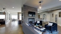 This thoughtfully designed single-floor space blends industrial and contemporary aesthetics to create a warm yet modern atmosphere. At the heart of the living area stands a dramatic floor-to-ceiling reclaimed wood accent wall, anchored by a sleek built-in fireplace and a mounted flat-screen television — serving as a striking focal point that draws the eye immediately upon entry. Polished concrete floors run throughout, adding an urban, loft-like quality that complements the exposed ductwork visible along the ceiling. A pendant light with a clear glass shade adds a refined touch overhead, balancing the raw industrial elements with a softer, inviting glow. The living area is furnished with plush gray sofas arranged around a clean-lined coffee table, creating a comfortable and stylish gathering space. Built-in shelving on one side of the accent wall provides functional display space. The open floor plan flows seamlessly into a dining area featuring bar-height seating, as well as an additional lounge or reading nook visible beyond the dividing accent wall. A separate shelving unit and bright, airy areas suggest generous natural light throughout. The overall ambiance is contemporary and welcoming, with a cohesive palette of gray, navy, and teal accents tying the space together.