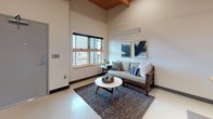This single-floor unit combines industrial and contemporary design elements throughout its well-organized layout. Distinctive exposed wood beam ceilings run the length of the space, adding warmth and architectural character to the otherwise clean, neutral interiors.

The living area features soaring ceiling height, a large wood-framed window that draws in natural light, smooth light-toned flooring, and a dedicated wall-mounted heating unit. The open layout flows naturally into the kitchen, which is outfitted with dark upper cabinetry, white lower cabinets, a white subway tile backsplash, stainless steel appliances including a full-size refrigerator, dishwasher, microwave, and electric range, complemented by a freestanding butcher block island beneath a pendant light fixture.

The bedroom is a calm, understated space with the same warm wood beam ceiling, a window with roller shade, a wall-mounted HVAC unit, and a sliding closet door providing ample storage. The bathroom offers a clean, functional design with a floating vanity, single undermount sink, and a full tub with gray tile surround enclosed by a curtain. Throughout the unit, the consistent use of neutral tones and natural materials creates a cohesive, modern ambiance.